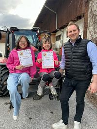 Zwei Kinder mit Urkunden sitzen vor einem Traktor, Mann steht daneben und lächelt
