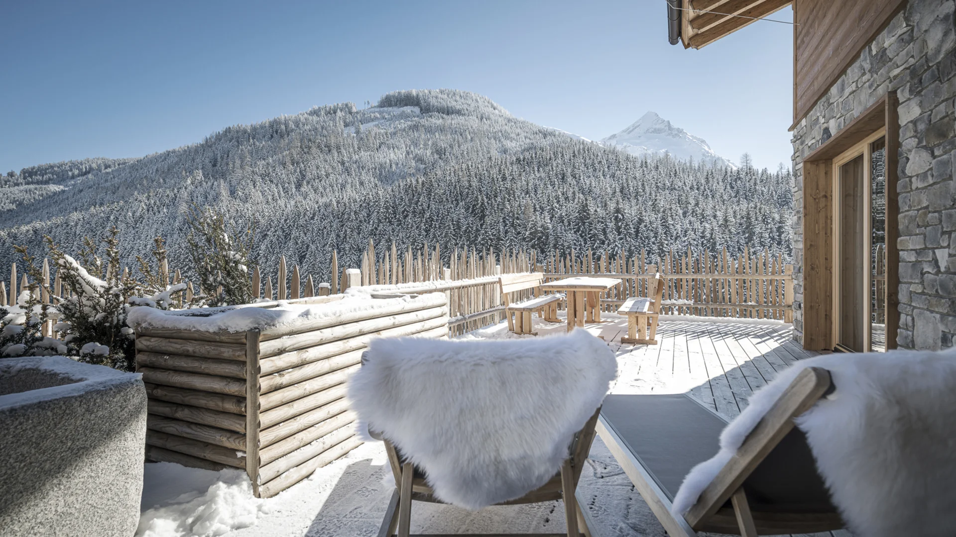 Farm Resort Geislerhof Winterterrasse mit Holzstühlen und Blick auf verschneite Berge und Wald